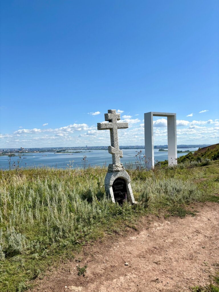 Крест и памятник на берегу реки, зеленые травы.